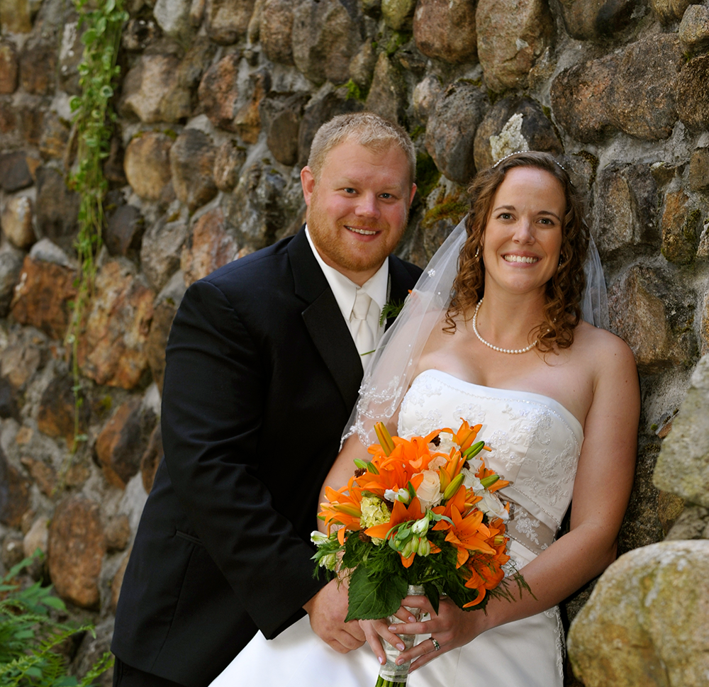 Man wearing a suit and woman wearing a white wedding dress standing against a brick background. 