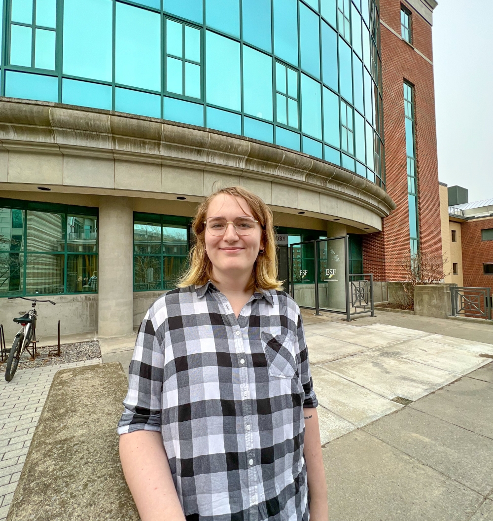 Woman in a plaid shirt standing in front of a building with large glass windows stone beams. 