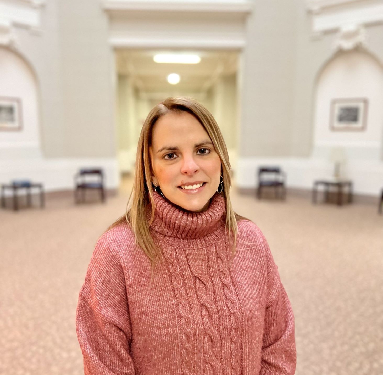 Woman in a pink sweater standing in a room with a pinkish brown carpet and tan and white walls. 