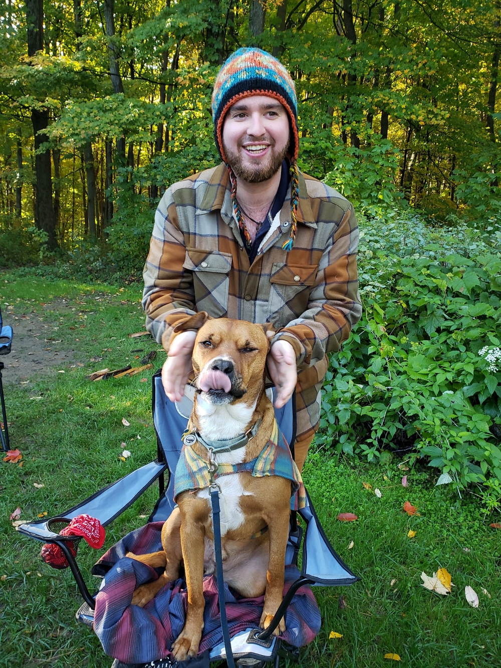 Man with a beanie hat and a flannel shirt standing in the middle of the woods. He has his hands around a brown and white dog. 
