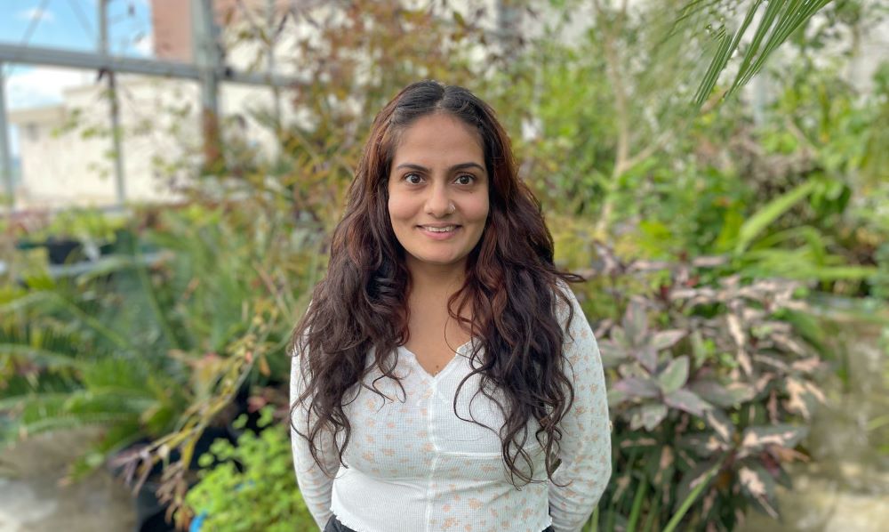 Woman with a white shirt standing with a background of green plants. 