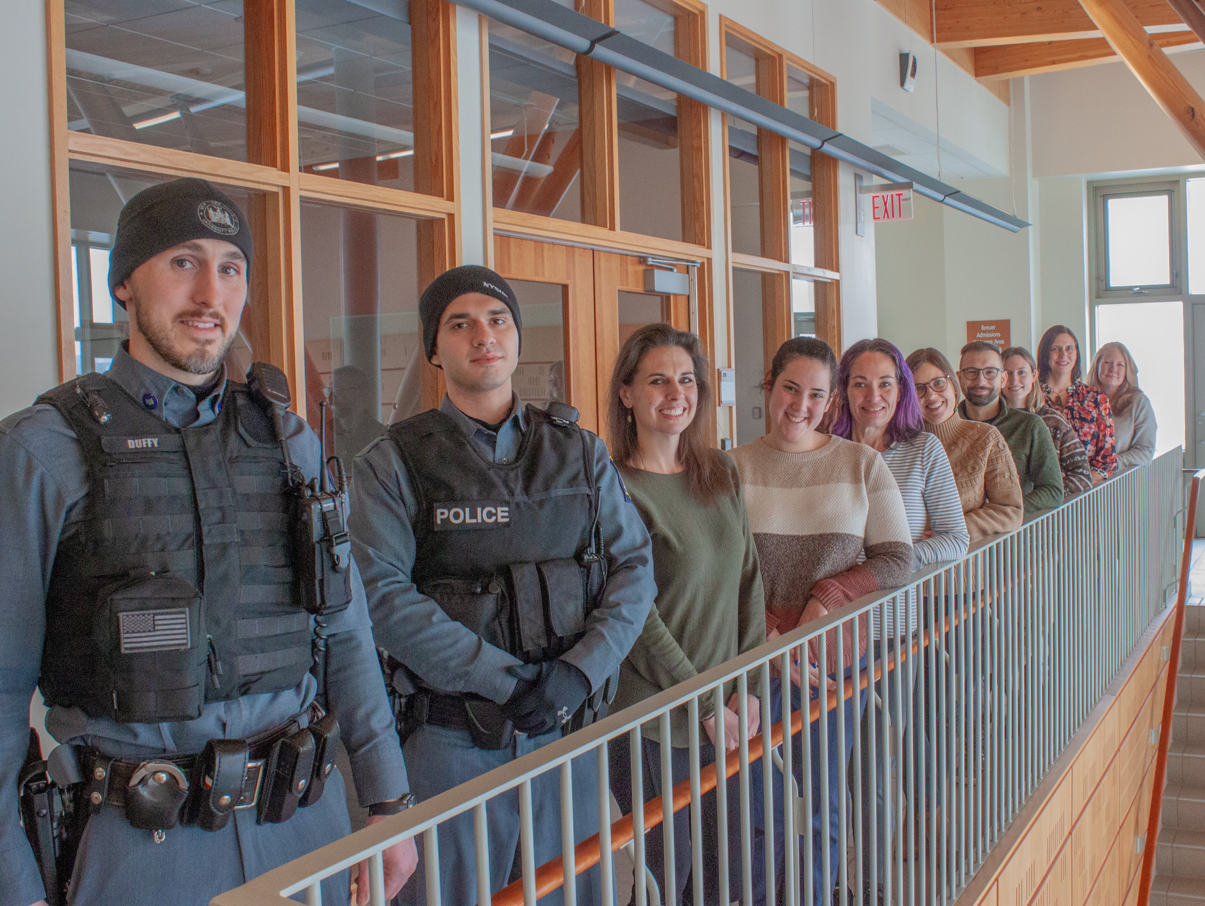 Row of people standing and looking at the camera. In the foreground is a brown railing and in the background is a room with glass you can see through. 