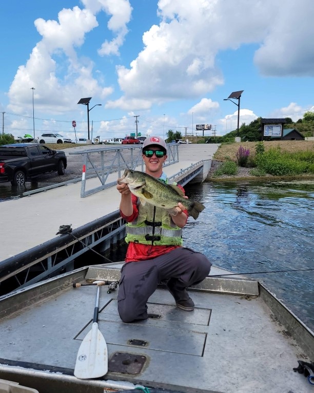 Man on a boat with a green life jacket, sunglasses and a white hat holding a fish. 