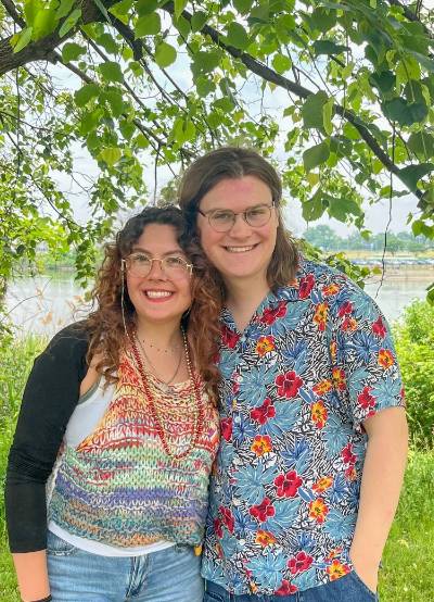 A man and a woman standing under a tree with a body of water in the background. They are both wearing multi-colored shirts! 