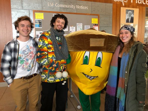 Three students standing near a giant yellow, green and brown acorn in front of a bulleting board. 