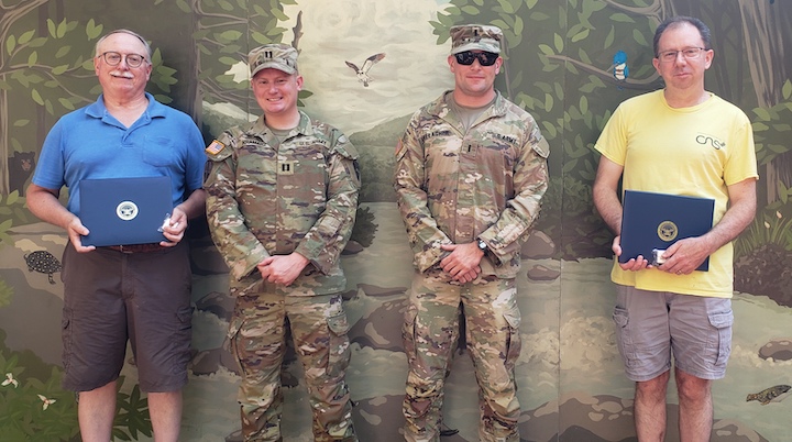 Four men standing in front of a mural. Man on left wearing blue shirt and shorts, two men in military uniforms and then a man in a yellow shirt and shorts.