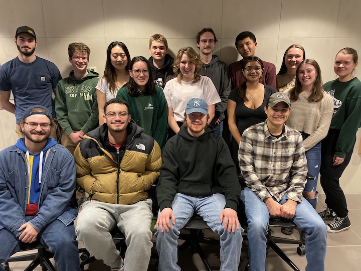 Sixteen students sitting for group photo against white wall.