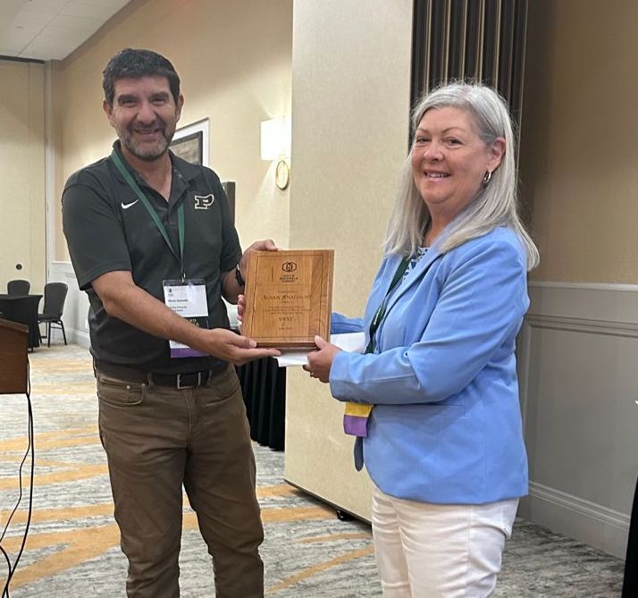 Man in green shirt and brown pants hands woman in blue jacket and white pants an award