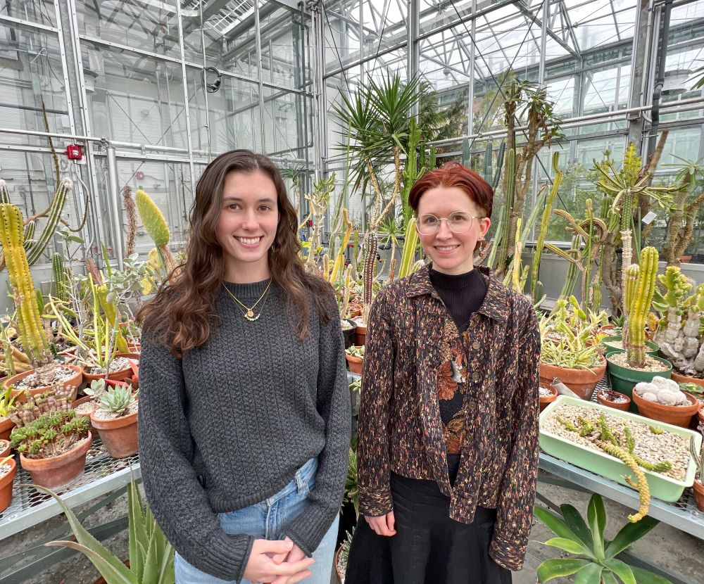 Two women, standing in a room full of cacti. They are wearing dark black colored clothing. 