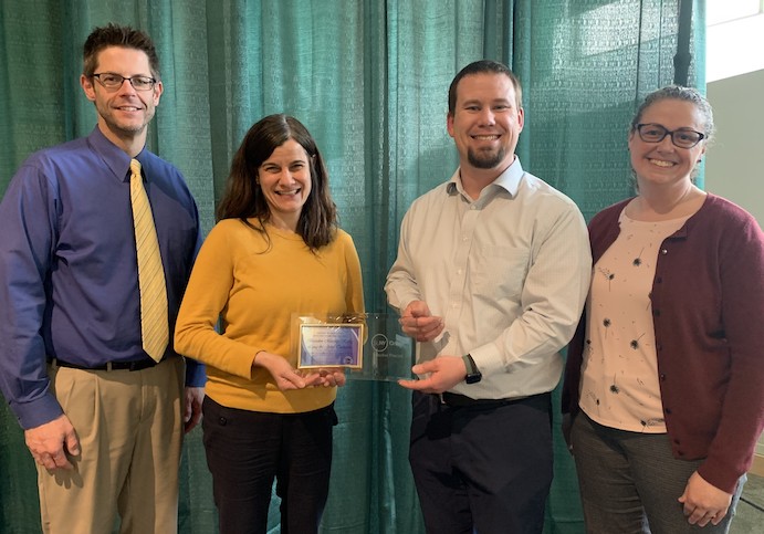 two men and two women standing by green curtain holding an award.