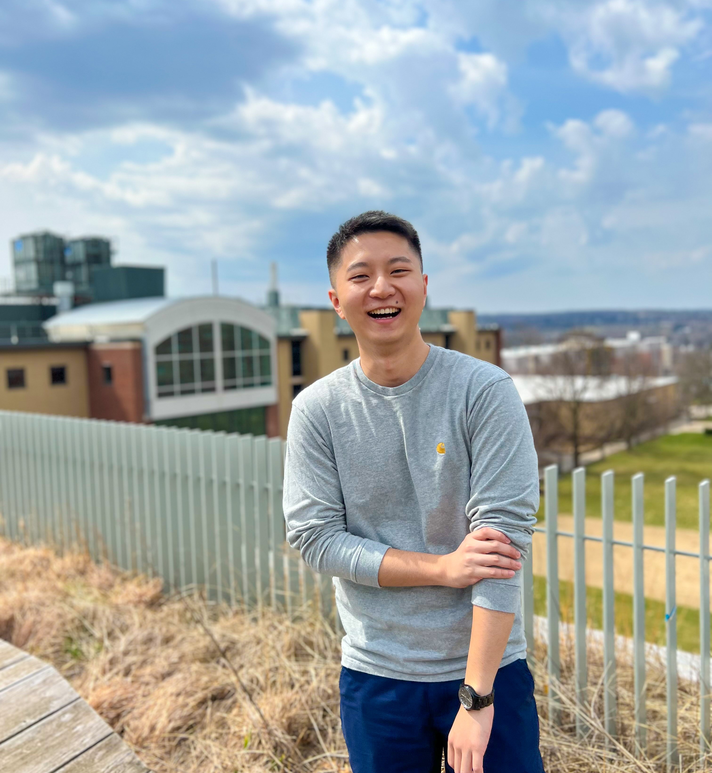 Man with a grey shirt standing outside with some buildings, a green field, and blue skies behind him. There is a fence that separates the horizon from the platform. 