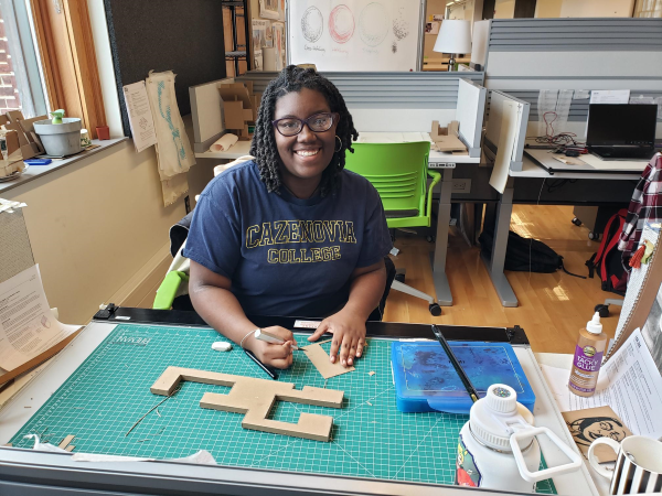 Woman wearing a Cazenovia College shirt and sitting at a desk with a bunch of design tools round her.