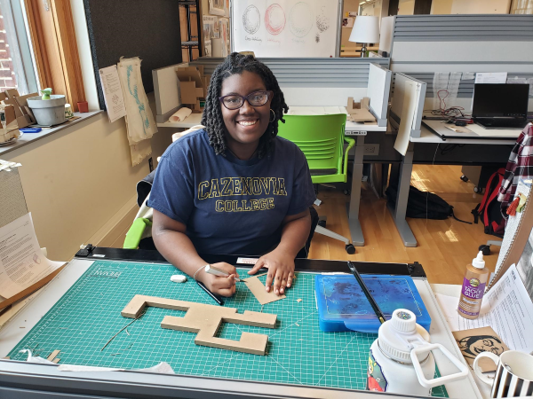 Woman wearing a Cazenovia College shirt and sitting at a desk with a bunch of design tools round her. 