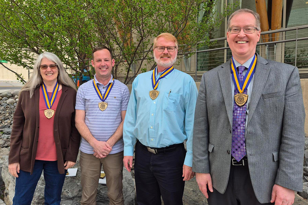 One woman and three men standing in front of a tree. All four are wearing medals on blue and gold ribbons.