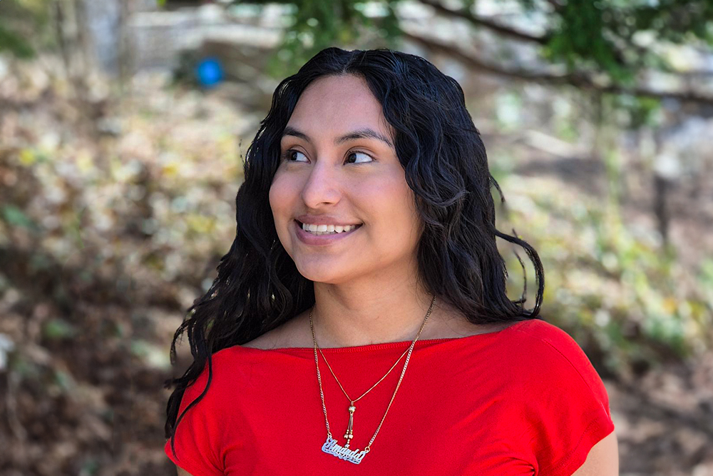 Young woman with long dark hair and red shirt smiling and looking to side. Standing outside.