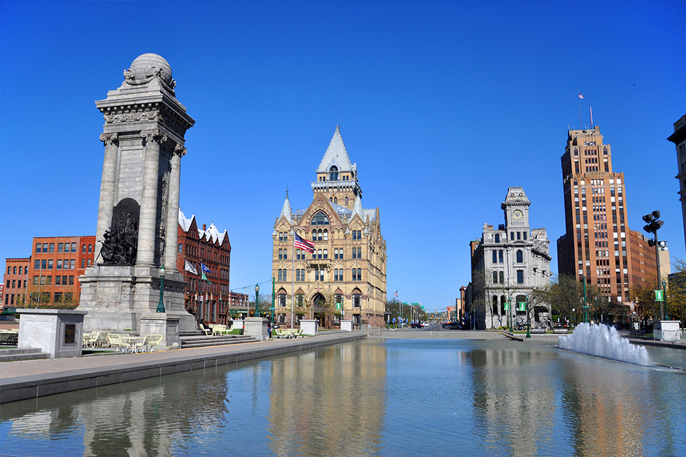 Large reflecting pool surrounded by three ornate buildings and one monument against a bright blue sky.