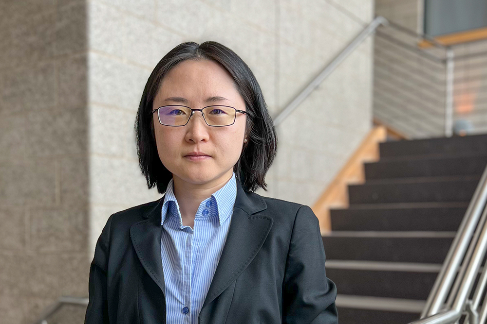 Woman with dark hair and glasses wearing blue shirt and suit jacket standing on stairs.