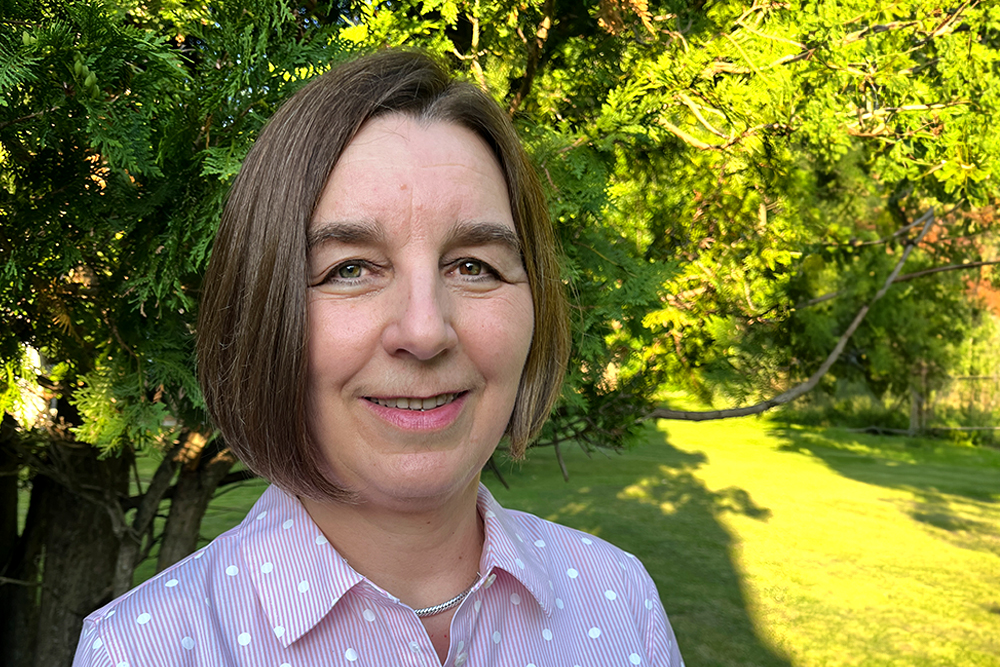 Woman with chin-length brown hair wearing striped pink shirt with polka dots standing in front of a tree.