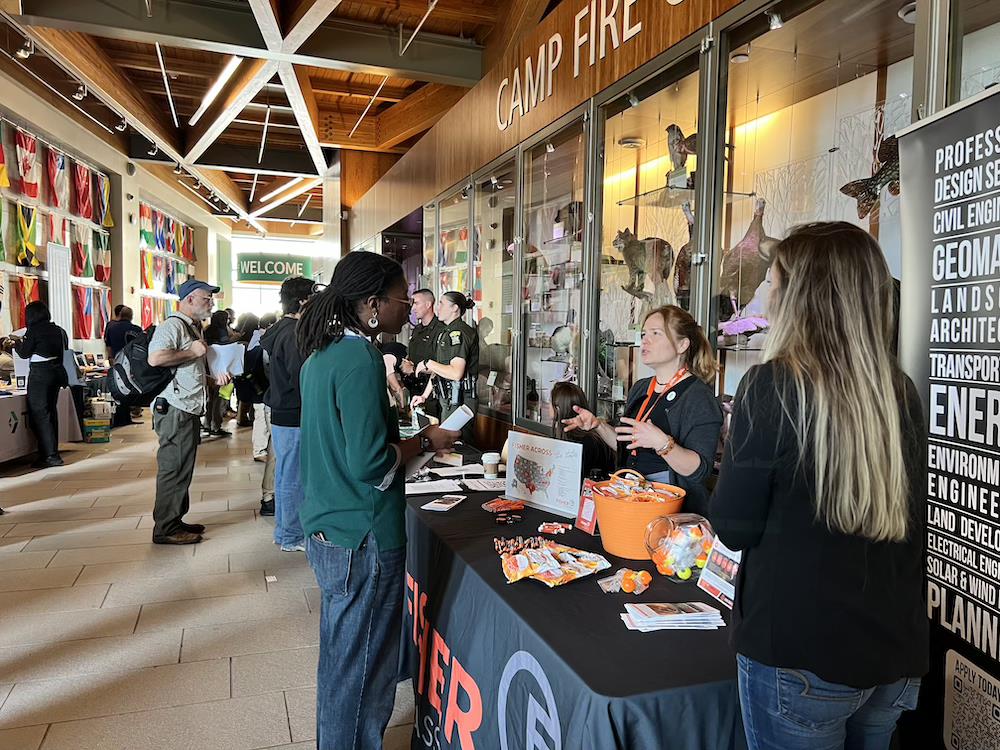 Young black woman in green shirt standing by a table talking with white woman wearing a blue shirt and orange lanyard.