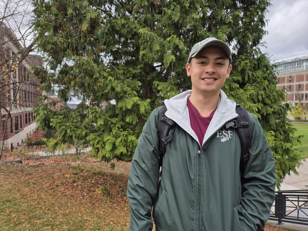 Man wearing baseball hat and green ESF-branded jacket standing in front of tree.