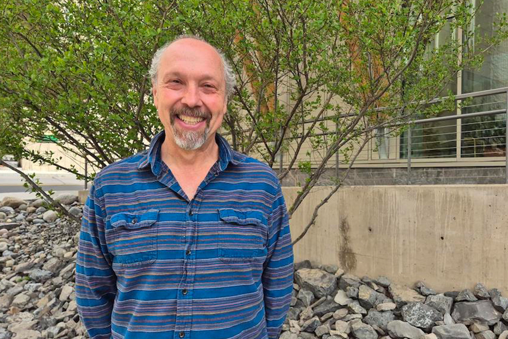 Man with goatee wearing a blue striped shirt standing in front of a tree.