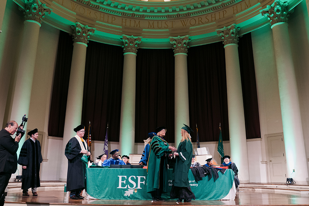 Two women wearing green graduation robes shaking hands in front of a green banner that says ESF.