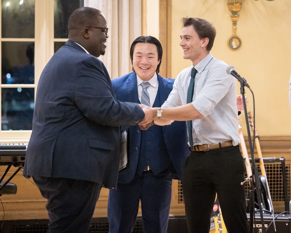 Two men smiling and shaking hands while third man looks on. 