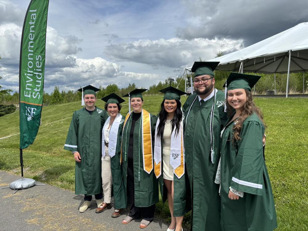 Six men and women wearing green graduation robes standing by banner that says Environmental Studies.