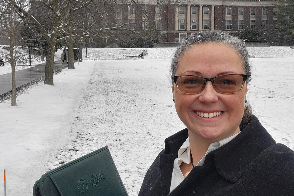 Woman in dark jacket and white shirt standing outside on snowy day in front of a brick building.