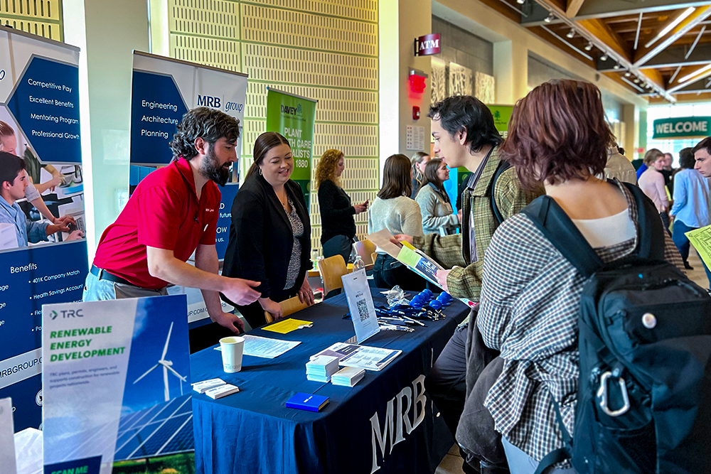 Man in red shirt talks with person in green shirt as they lean over a table at a career fair.