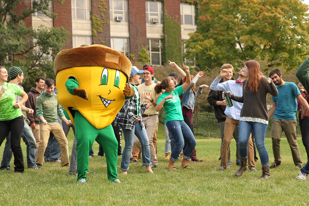 Oakie Acorn, large yellow acorn mascot, dances with students outside a brick building.