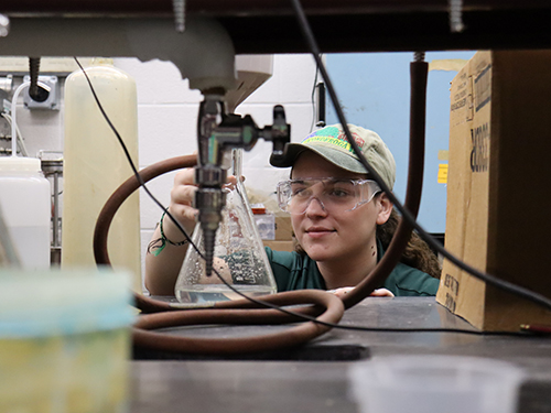 Nicole Byrnes working in the lab. She is wearing safety glasses and holding a conical flask