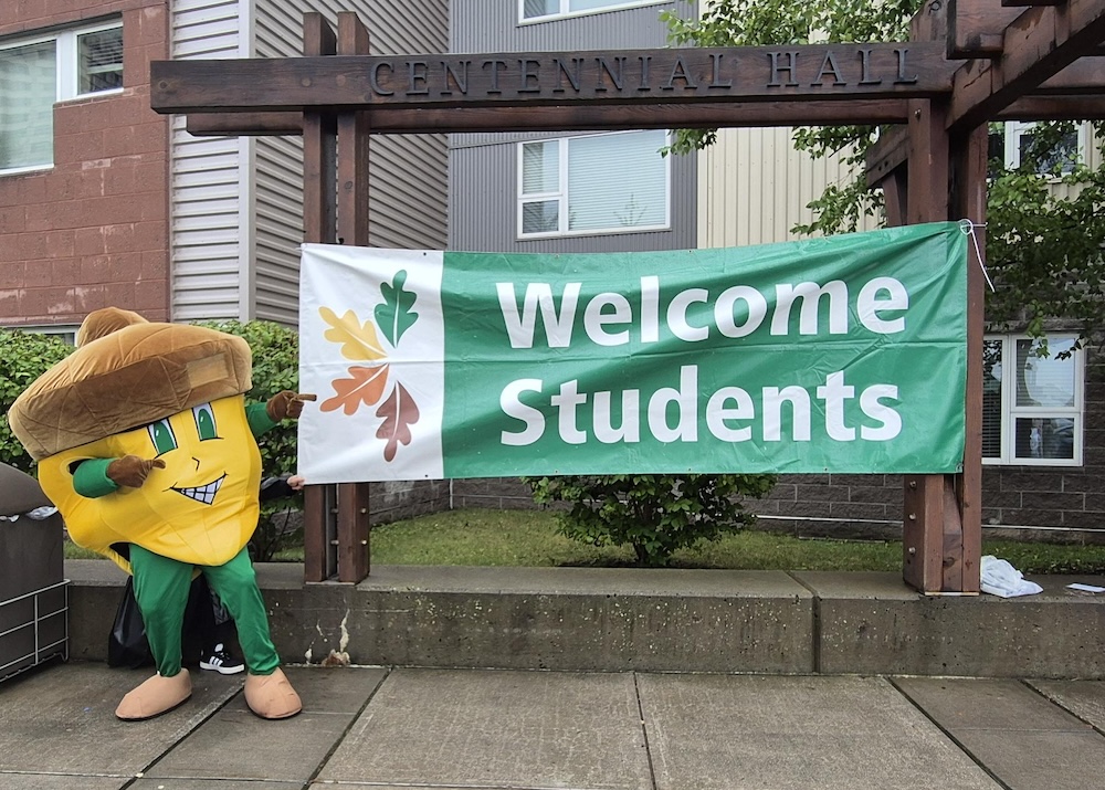 Giant acorn mascot with smiling face in front of green Welcome Students sign.