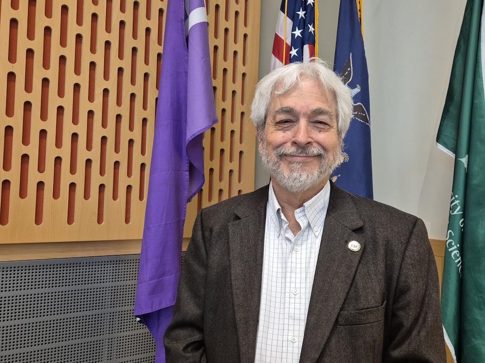 Man with white hair, beard and mustache standing in front of flags.