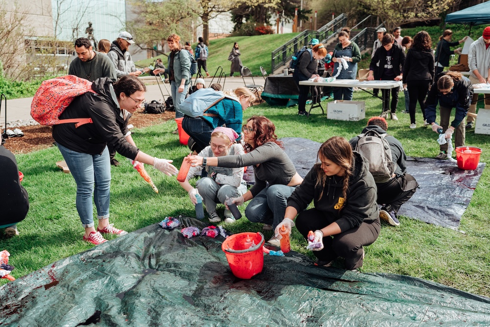 Large group of students tie-dying shirts using bottles of dye while gathered around a tarp.