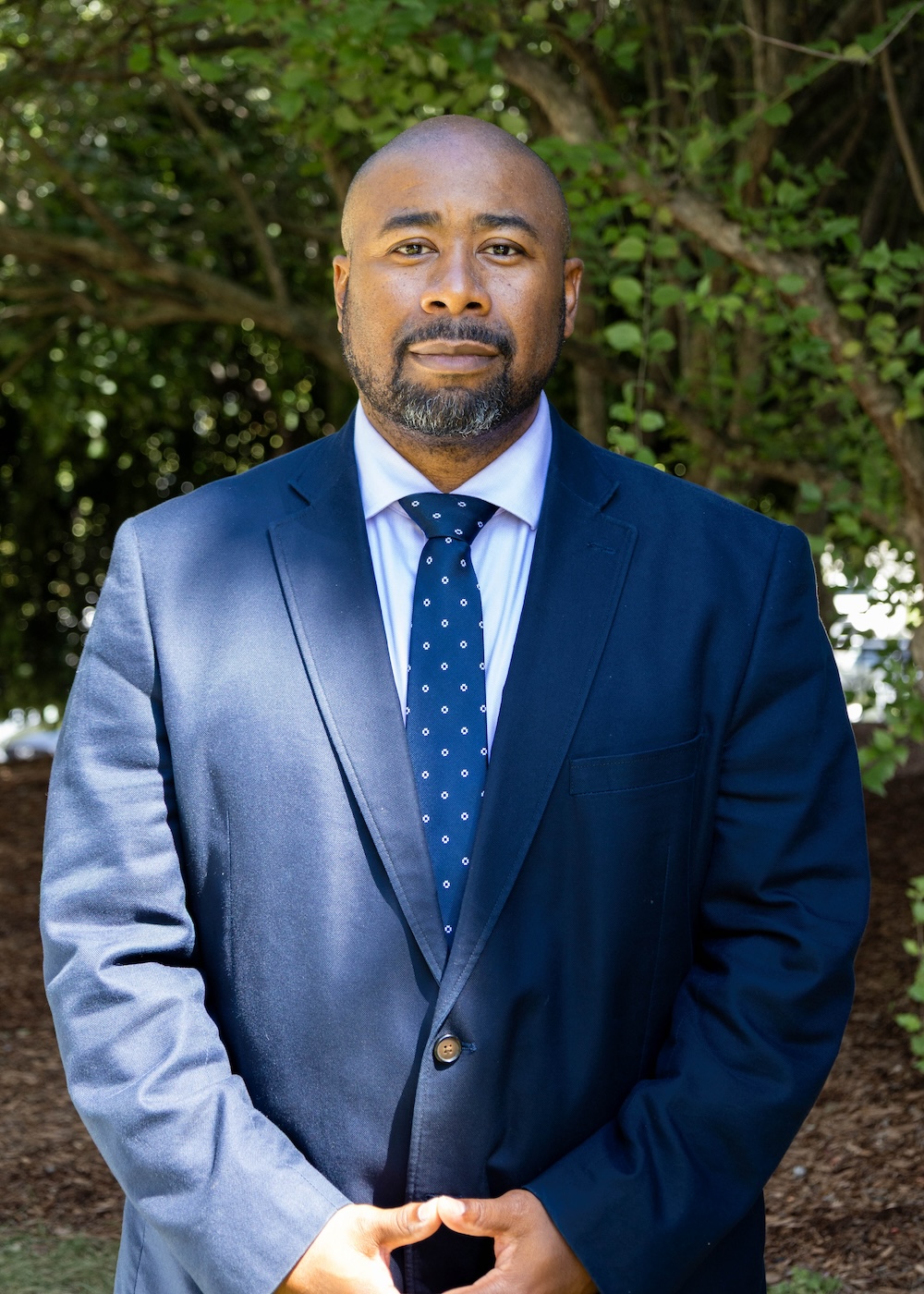 Black man with beard and mustache wearing a blue suit stands in front of a greenery.