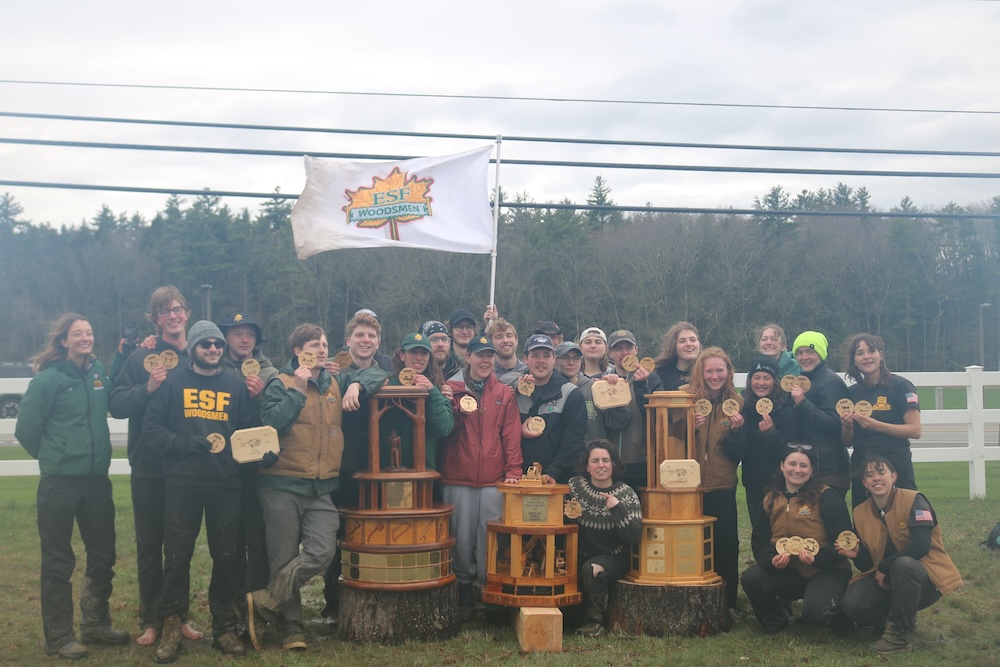 Group of young men and women posing with three large trophies and a flag with a yellow maple leaf.