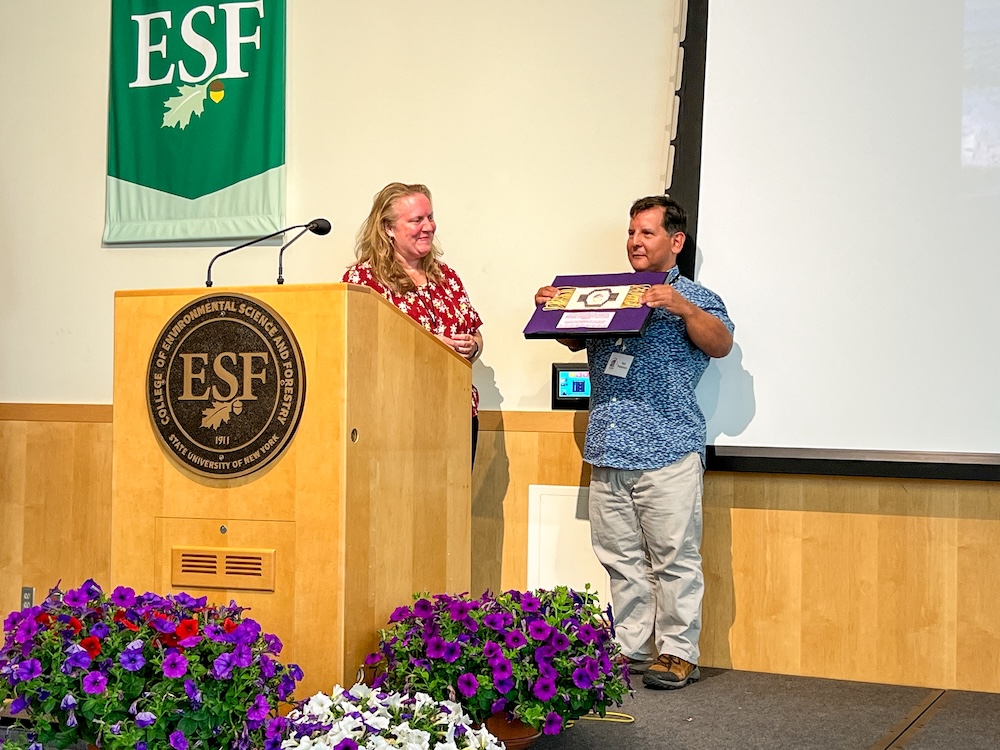 A speaker stands at a wooden podium with an ESF emblem while another person stands beside the stage area holding up a framed item for display. Purple flowers line the front of the stage, and an ESF banner and projection screen are visible in the background.