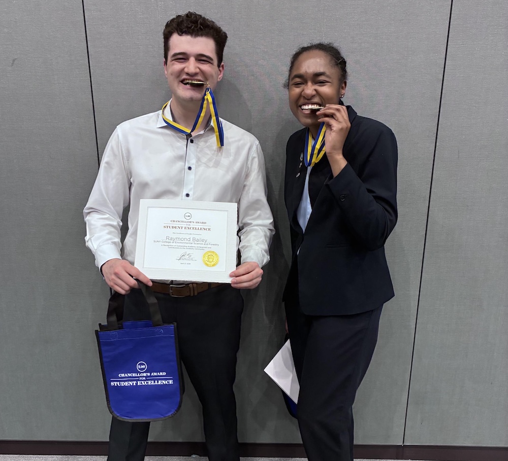 Two people stand against a gray wall, each holding a medal ribbon near their mouth. One person also holds a framed Student Excellence certificate and a blue bag with matching text.
