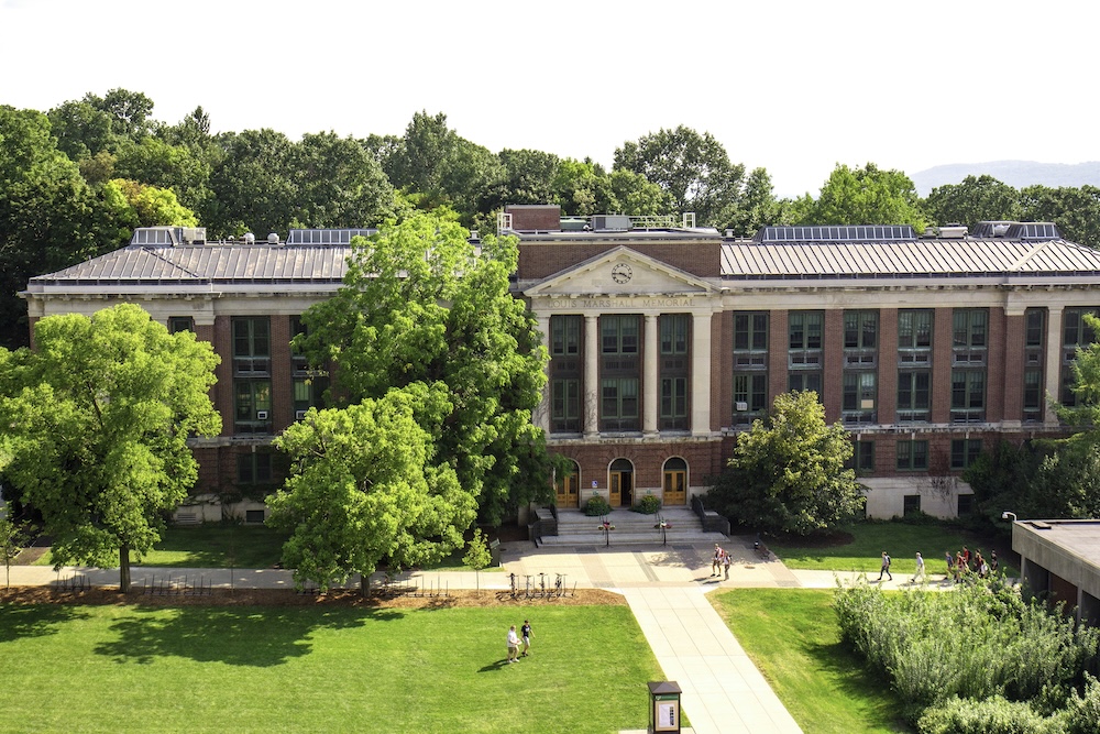 A large, historic brick academic building with a columned central entrance sits behind a landscaped lawn and broad pedestrian walkway. Mature green trees frame the building, while several people walk across the campus grounds on a sunny day.