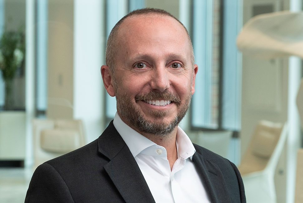 A professional head-and-shoulders portrait shows a person wearing a dark blazer over a light-colored collared shirt, standing indoors. The background features large windows and softly blurred modern seating elements, creating a bright, contemporary setting.