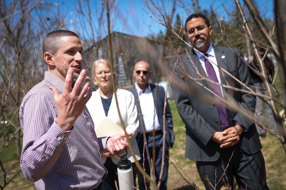 A small group of people stands outdoors among young, leafless trees on a sunny day, with one person in the foreground gesturing while speaking. Others stand nearby listening, with a building and grassy area visible in the background.