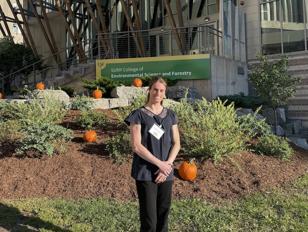A person wearing a dark top and black pants stands in front of the SUNY College of Environmental Science and Forestry sign outside a campus building. The landscaped area around the sign is decorated with several orange pumpkins, and the building’s distinctive wood-and-glass architectural features are visible in the background.