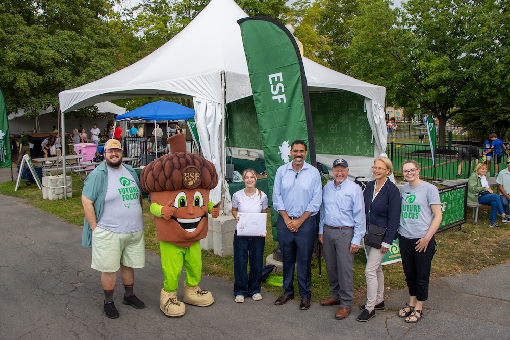 A group of people and a large acorn mascot stand in front of an ESF-branded tent at an outdoor fairground event. Visible signage includes “ESF” and “Future Focus,” with additional tents, visitors, trees, and activity areas in the background.