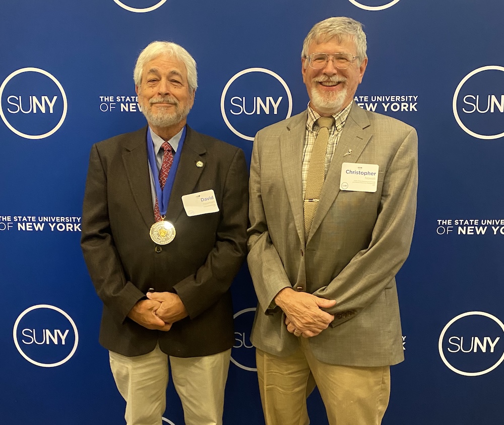 Two individuals stand in front of a blue backdrop displaying SUNY and State University of New York logos. One individual wears a medal over a suit jacket, and both wear name badges while posing for a photo at a recognition event.