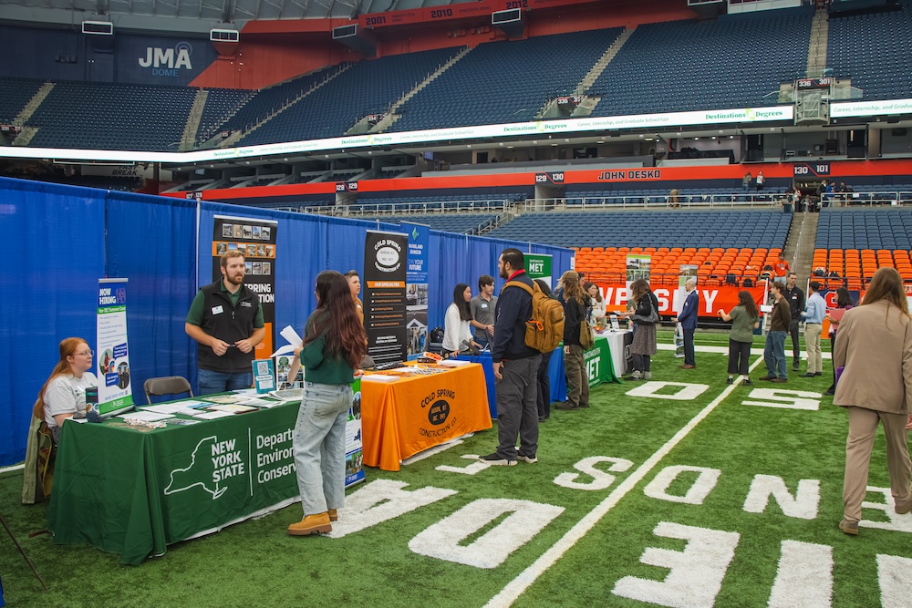 a career and graduate school fair taking place on the turf inside the JMA Dome, with employer and organization booths arranged in rows as attendees speak with recruiters and gather information. In the background, stadium seating and signage are visible, highlighting the large-scale indoor venue hosting the networking event.