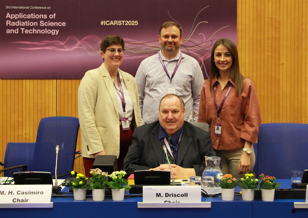 Four adults pose at a conference table. One person sits centered at the table wearing a dark suit and tie, smiling toward the camera, while three colleagues — two standing behind on the left and center and one standing on the right —s tand close together, all wearing conference lanyards and smiling. The table in front has microphones, a water pitcher and glasses, small potted plants, and name placards indicating chair roles. Behind them, a large banner reads “3rd International Conference on Applications of Radiation Science and Technology” with the hashtag “#ICARST2025.” 