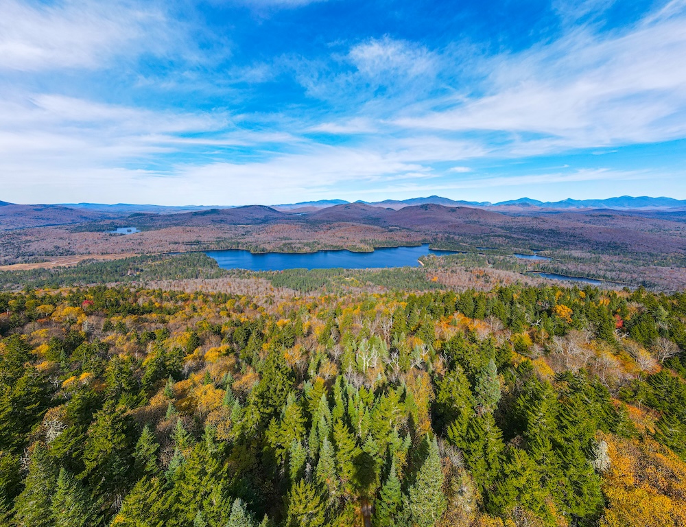 Drone view of mountains with trees in the foreground, and a lake and more mountains in the background.