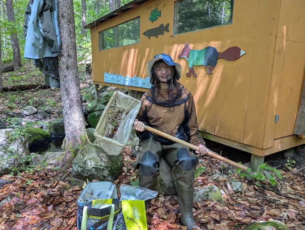 A person wearing waterproof field gear sits in a forested area holding a large rectangular sampling net beside bags of equipment. Behind the person is a small wooden structure decorated with colorful animal cutouts, surrounded by rocks, trees, and a nearby stream.