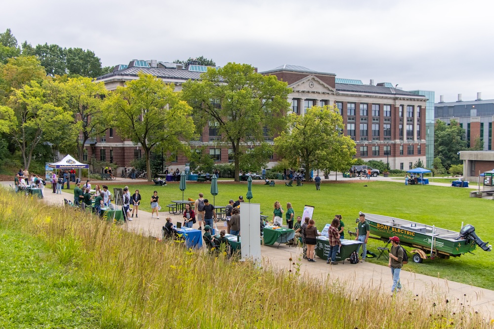 Students and community members gather at outdoor information tables and displays on a campus lawn, with a historic brick academic building and mature trees in the background. A grassy meadow in the foreground overlooks the event, where attendees explore exhibits, talk with exhibitors, and view a small electric boat on display.